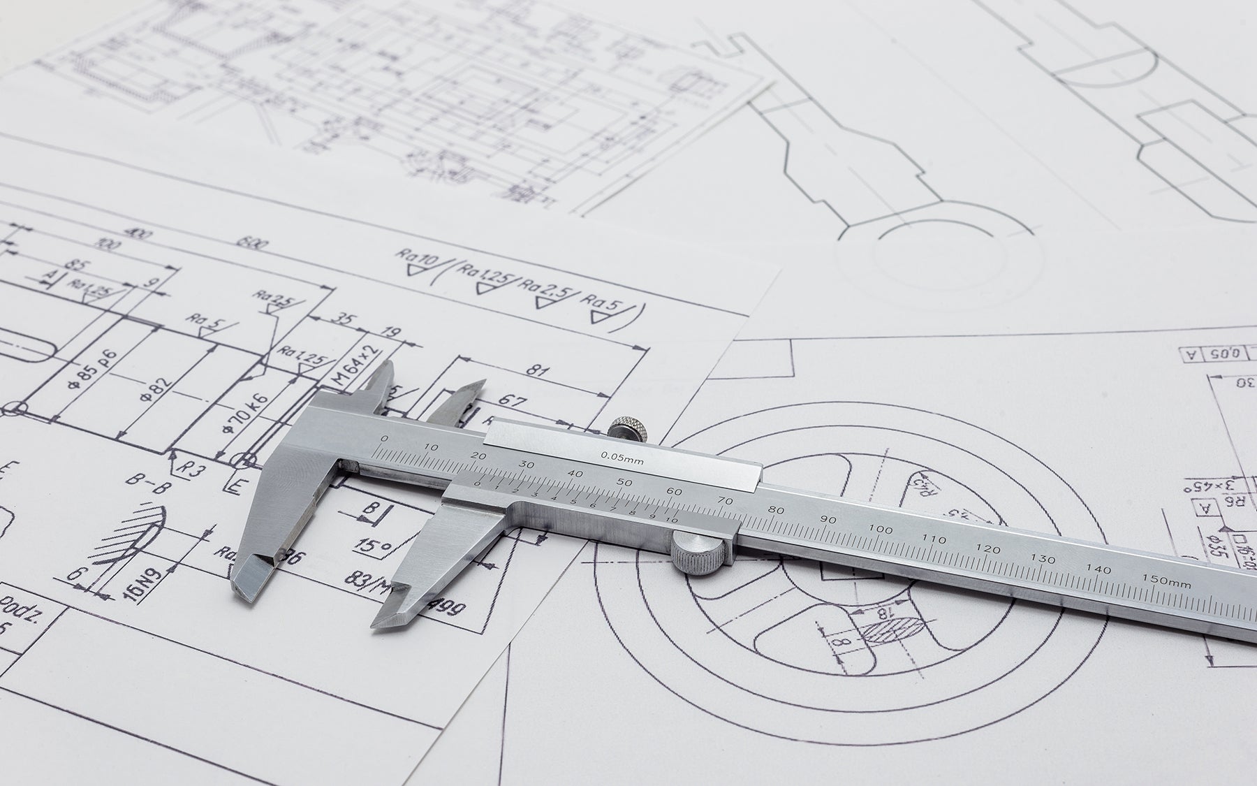 A metal caliper rests on several technical engineering drawings with detailed measurements and schematics on white paper.